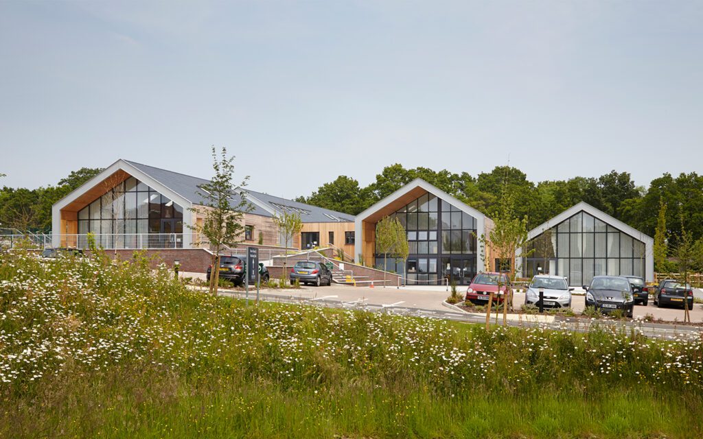 Three large barn-style school buildings with large glazed front, pitched roofs, and cars parking in front.