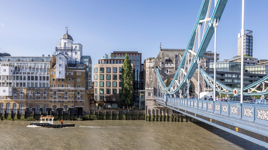 Brick and metal cladded building with reflective windows with the River Thames and Tower Bridge in front.
