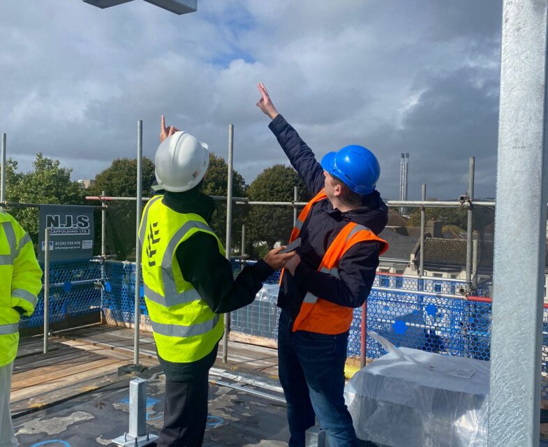 Two architectural technologists wearing high-vis vests and hard hats point to a construction detail on a site visit.