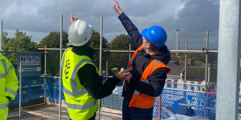 Two architectural technologists wearing high-vis vests and hard hats point to a construction detail on a site visit.