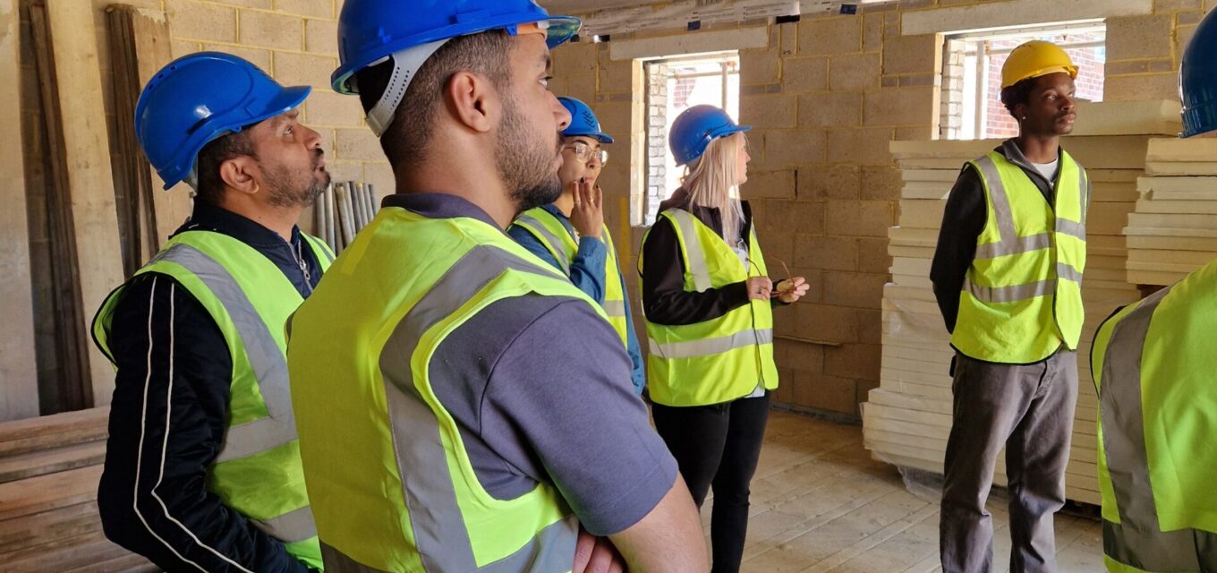 Group of five architectural staff wearing high-vis vests and hard hats looking at an unfinished building.