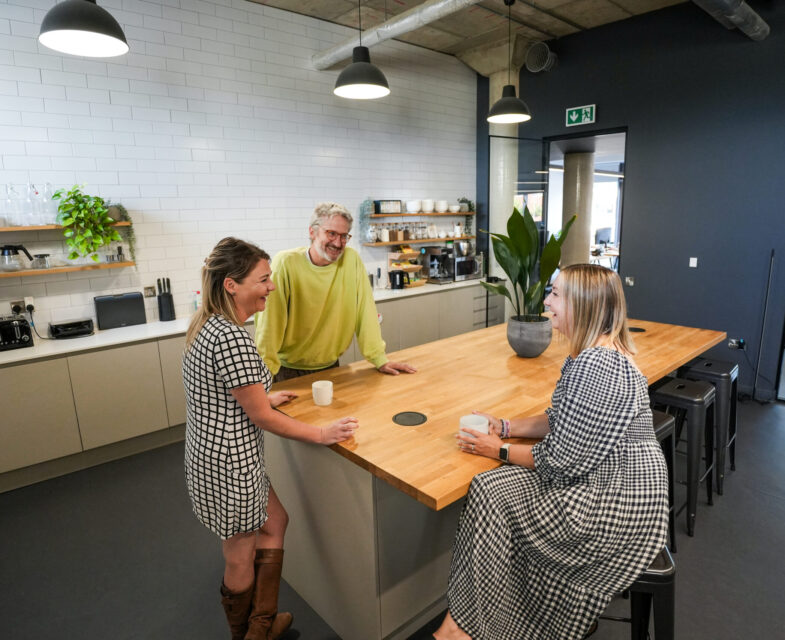 Three people sitting and standing around a kitchen island talking to each other and smiling.