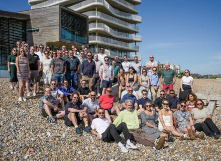 Large group of people on the beach in front of a tall residential building, all looking at the camera.