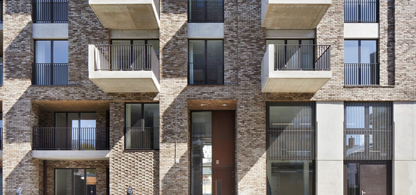 Grey brick building with large windows and protruding balconies with concrete bases and metal railing.