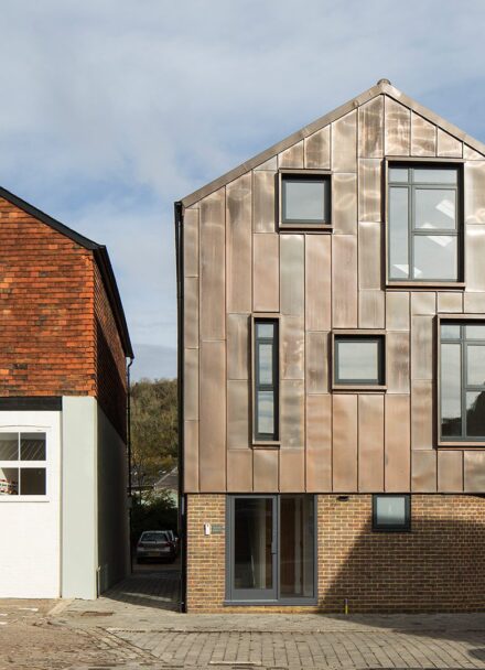 Contemporary residential building with metal cladding and a curved bottom wall sitting next to a traditional house with the same roof line.