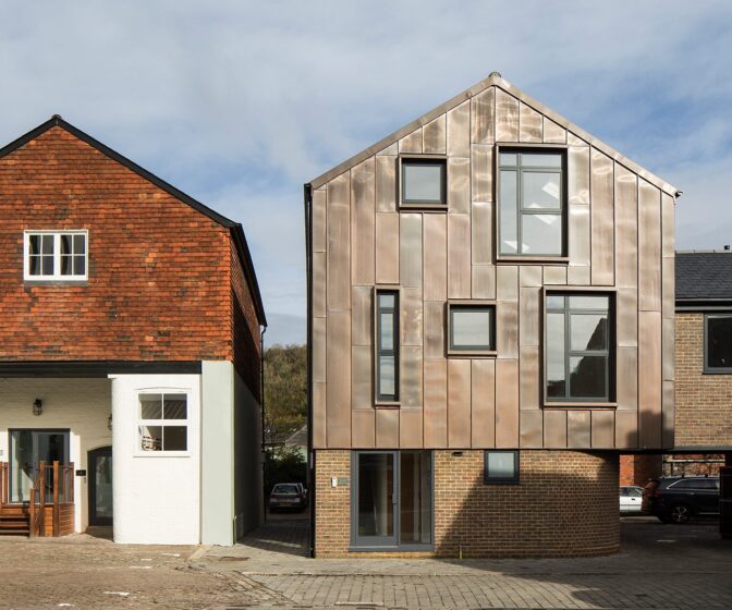 Contemporary residential building with metal cladding and a curved bottom wall sitting next to a traditional house with the same roof line.