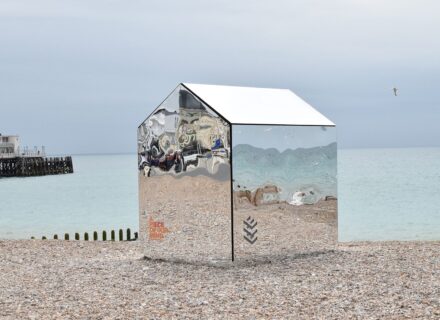 A beach hut made entirely from mirrors, standing on a pebble beach with a pier and the sea in the background.
