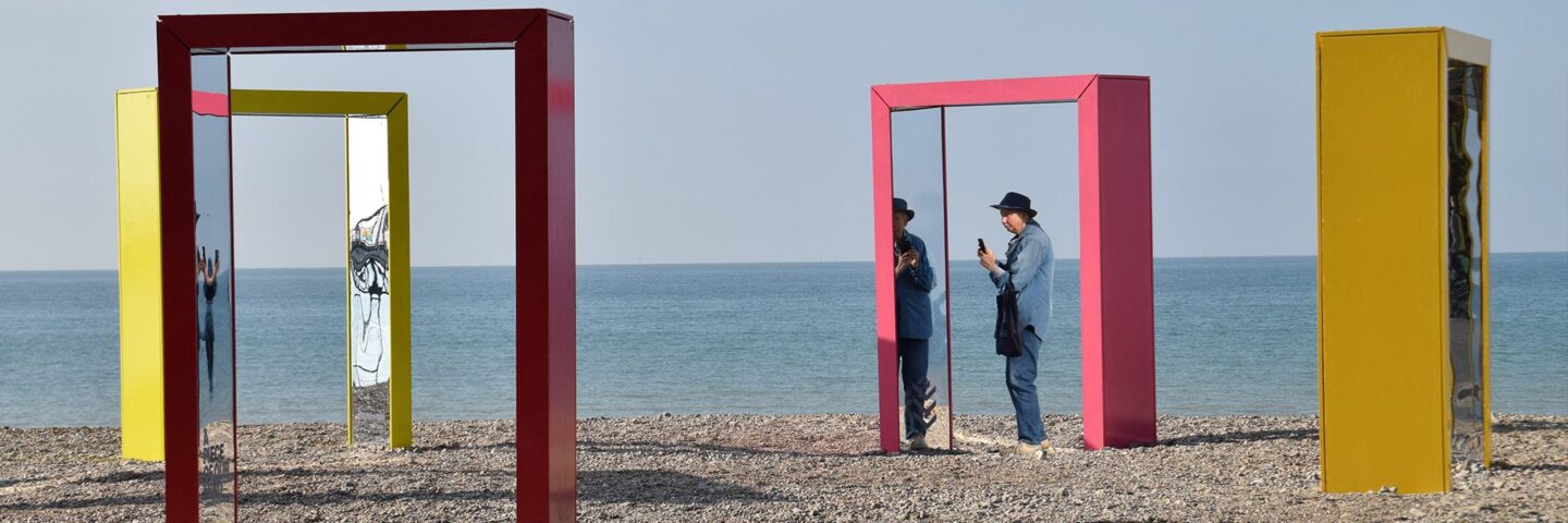 Four colourful archways with mirrors standing on a beach. A person is standing under one of them, taking a picture.