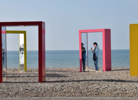 Four colourful archways with mirrors standing on a beach. A person is standing under one of them, taking a picture.