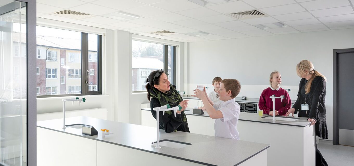 Science class room with three students and two teachers standing by tall desks with sinks, white walls and large windows.