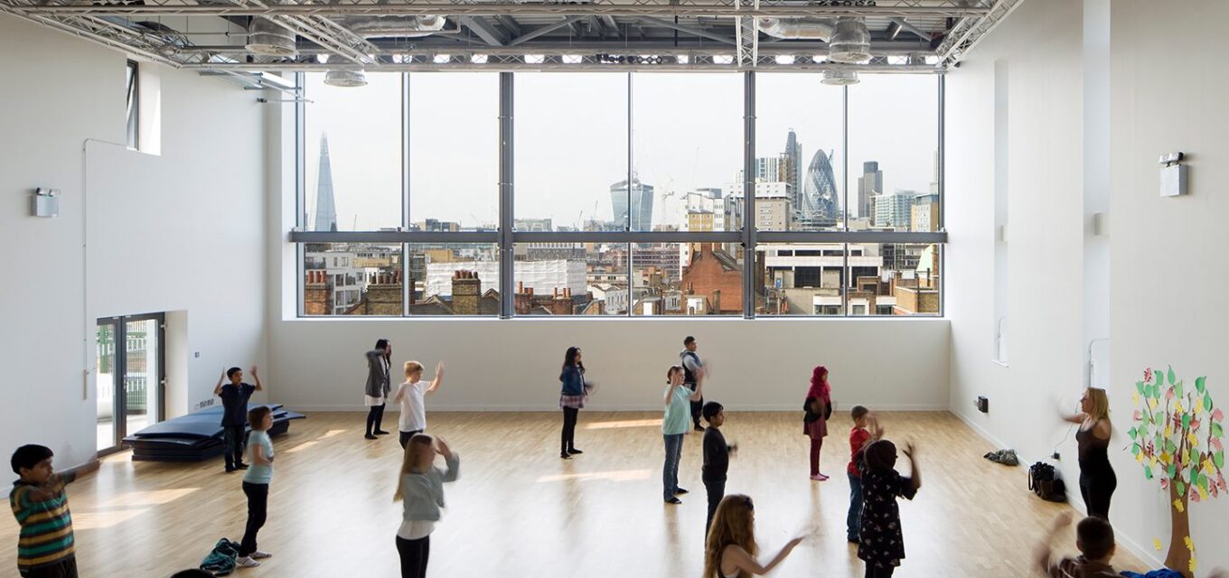 Large open dance studio with children and a teacher spread throughout the space. Large windows to the back overlook the London skyline with the Shard.