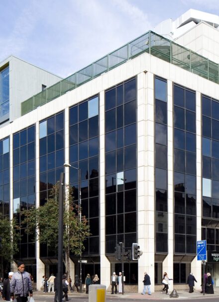 Modern, square building with large blackened glazing fronts next to high street filled with people shopping.