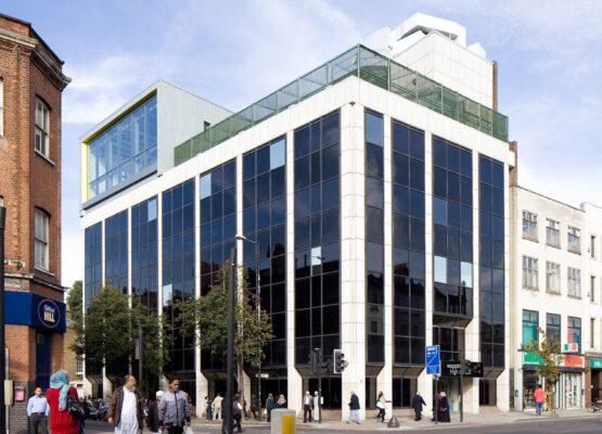 Modern, square building with large blackened glazing fronts next to high street filled with people shopping.