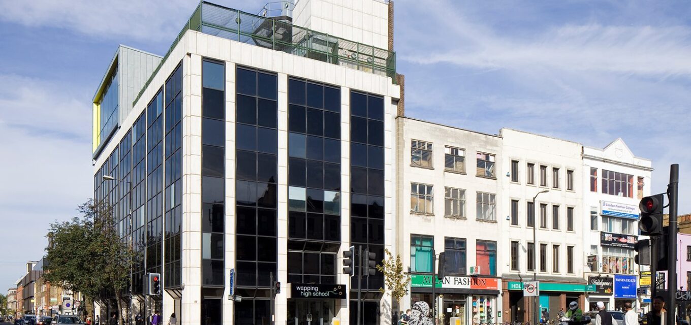 Large modern building with blackened windows next to high street shops filled with people shopping.