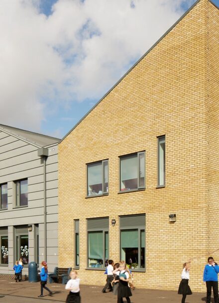 Primary school building with four different sections and angled roof lines. Three of the sections have grey cladding with the main building covered in yellow brick. School children are playing in front of the building.
