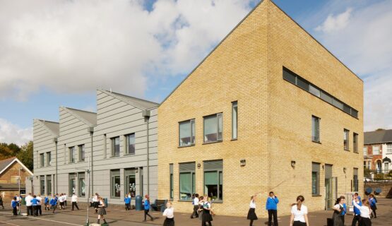 Primary school building with four different sections and angled roof lines. Three of the sections have grey cladding with the main building covered in yellow brick. School children are playing in front of the building.