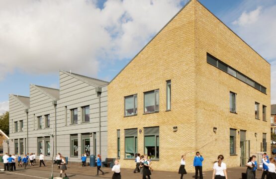 Primary school building with four different sections and angled roof lines. Three of the sections have grey cladding with the main building covered in yellow brick. School children are playing in front of the building.