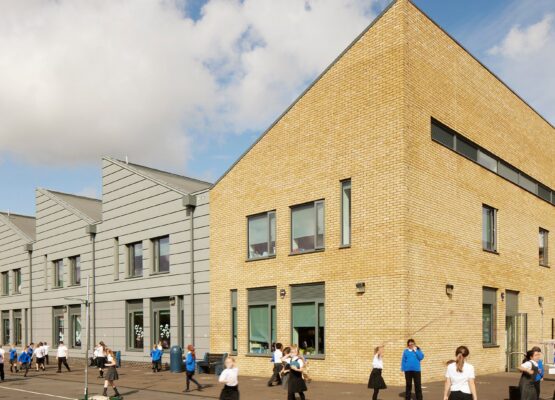 Primary school building with four different sections and angled roof lines. Three of the sections have grey cladding with the main building covered in yellow brick. School children are playing in front of the building.