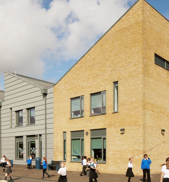 Primary school building with four different sections and angled roof lines. Three of the sections have grey cladding with the main building covered in yellow brick. School children are playing in front of the building.