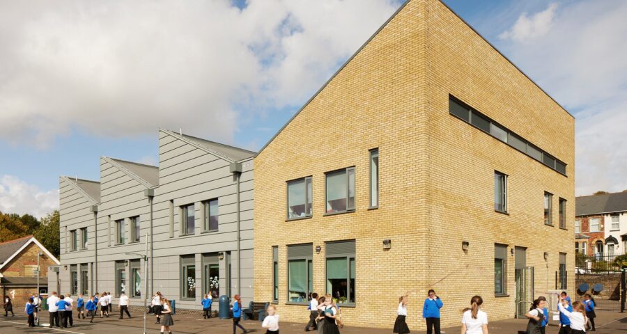 Primary school building with four different sections and angled roof lines. Three of the sections have grey cladding with the main building covered in yellow brick. School children are playing in front of the building.