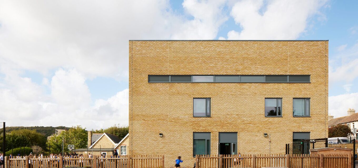 Square yellow brick school building with long window at the top behind a wooden picked fence and children running.