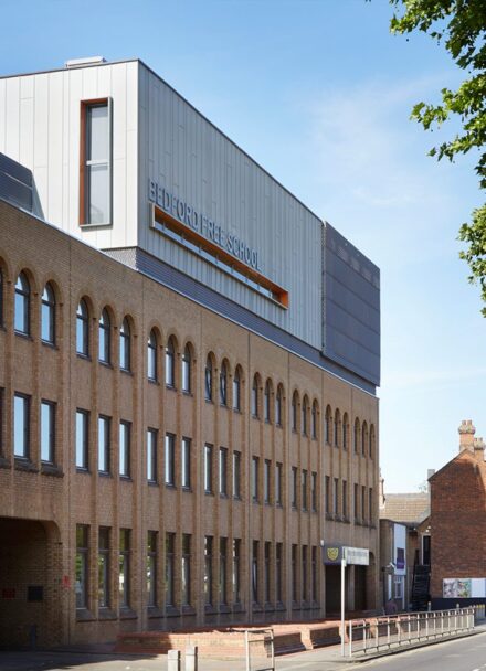 Brick clad school building with metal clad roof extension looking like a box with orange-detail windows.