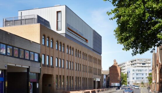 Brick clad school building with metal clad roof extension looking like a box with orange-detail windows.