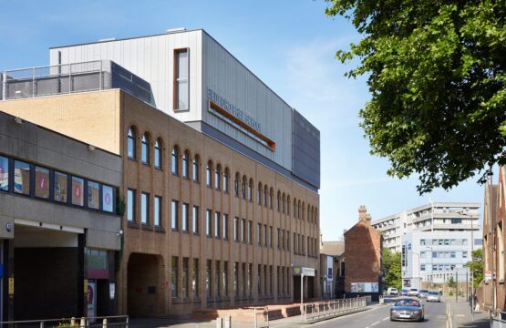 Brick clad school building with metal clad roof extension looking like a box with orange-detail windows.