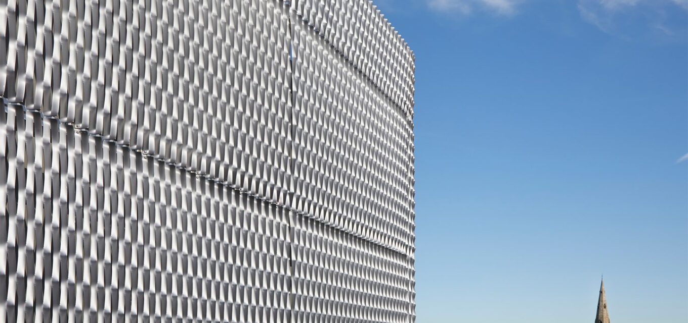 Metal mesh cladding on school building overlooking church tower and trees.