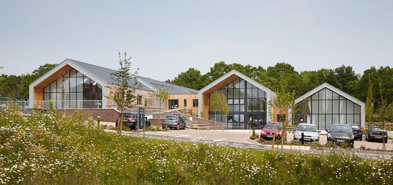 Three large barn-style school buildings with large glazed front, pitched roofs, and cars parking in front.