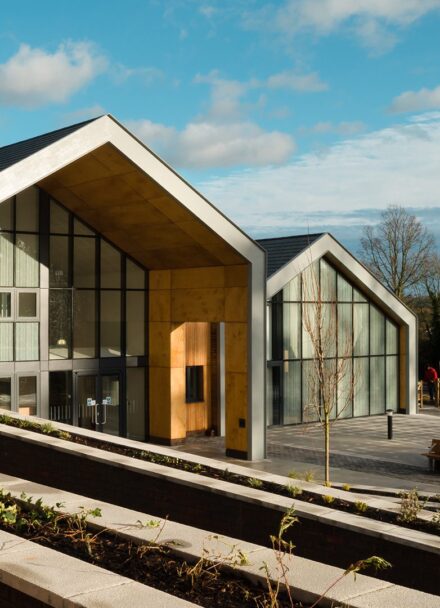 Two large school buildings in a barn-style with large glazed fronts and paved front area with trees and plants in a rural setting
