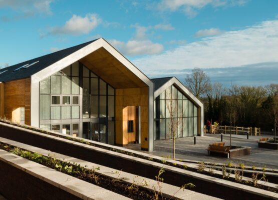 Two large school buildings in a barn-style with large glazed fronts and paved front area with trees and plants in a rural setting