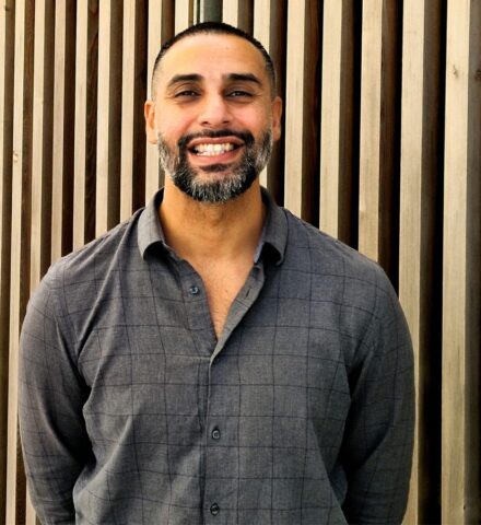 Person standing in front of timber clad building, smiling at the camera.