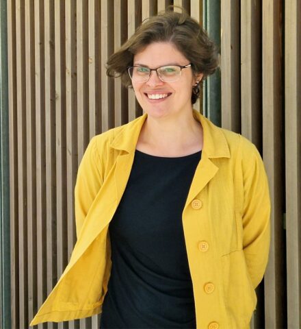 Person standing in front of a timber clad building, smiling at the camera.
