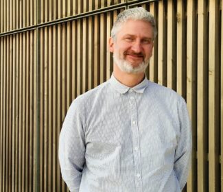 Person standing in front of a timber-cladded building, smiling at the camera.