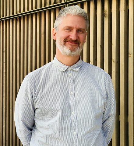 Person standing in front of a timber-cladded building, smiling at the camera.