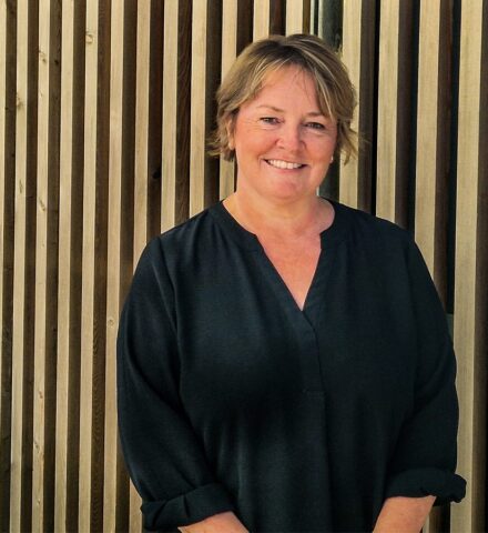 Person standing in front of timber cladded building, smiling at the camera.