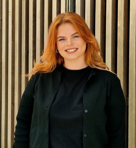 Person standing in front of a timber clad building, smiling at the camera.