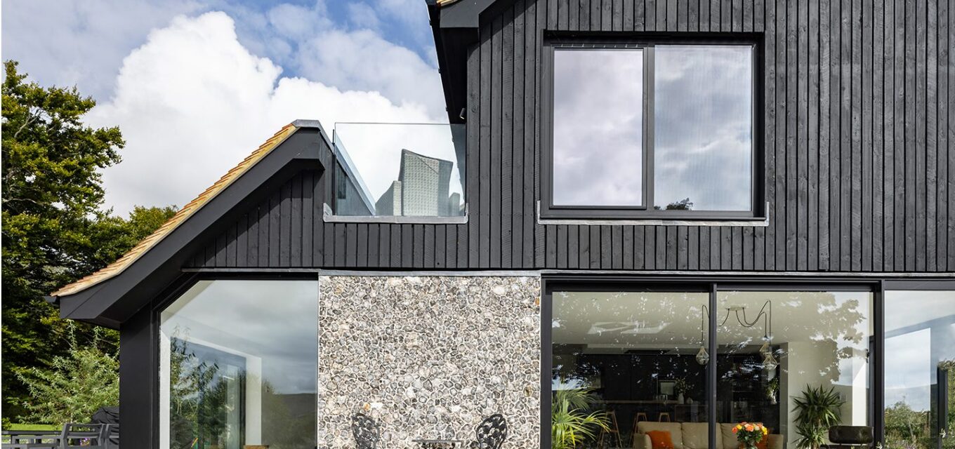 Stone terrace of a residential home with flint walls, black wooden cladding and continuous roof line with a glass balustrade balcony.