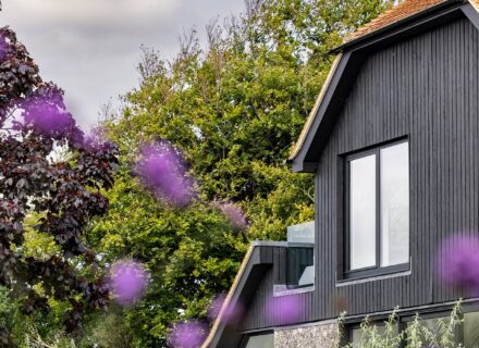 Private house with black wooden cladding and large windows with blurred purple flowers in the front.
