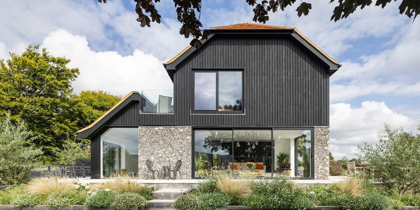 Barn-style residential house with flint walls, black wooden cladding and red tiled roof with plants in front.
