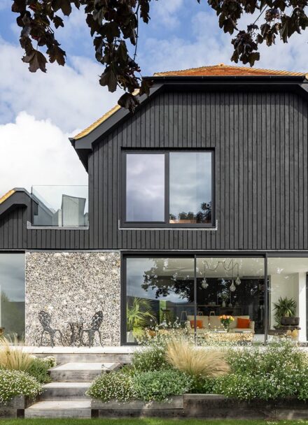 Barn-style residential house with flint walls, black wooden cladding and red tiled roof with plants in front.