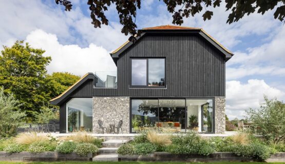 Barn-style residential house with flint walls, black wooden cladding and red tiled roof with plants in front.