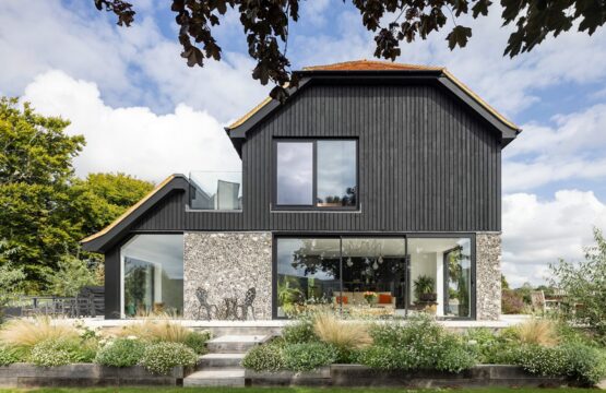 Barn-style residential house with flint walls, black wooden cladding and red tiled roof with plants in front.