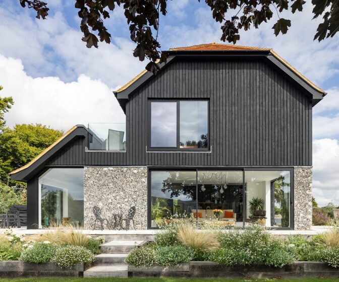 Barn-style residential house with flint walls, black wooden cladding and red tiled roof with plants in front.