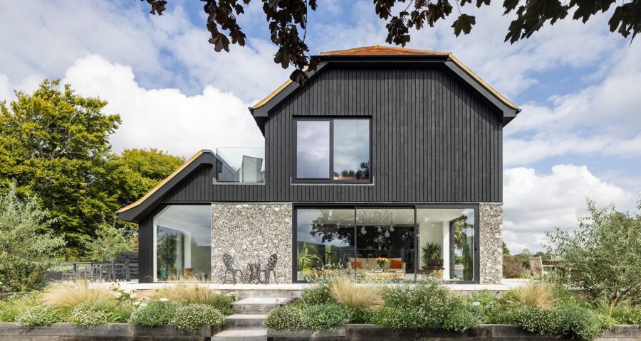 Barn-style residential house with flint walls, black wooden cladding and red tiled roof with plants in front.