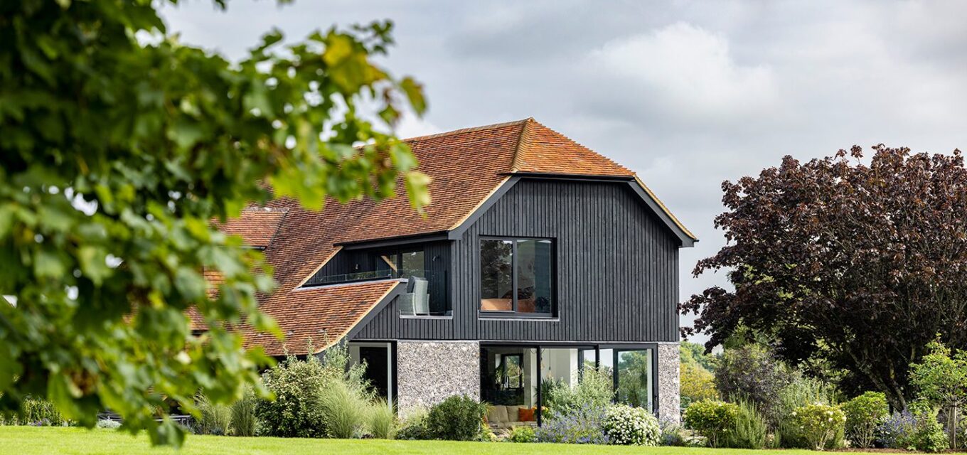 Barn-style residential house with large windows, black wooden cladding and flint walls in a garden with flowers.