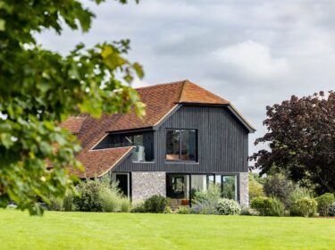 Barn-style residential house with large windows, black wooden cladding and flint walls in a garden with flowers.