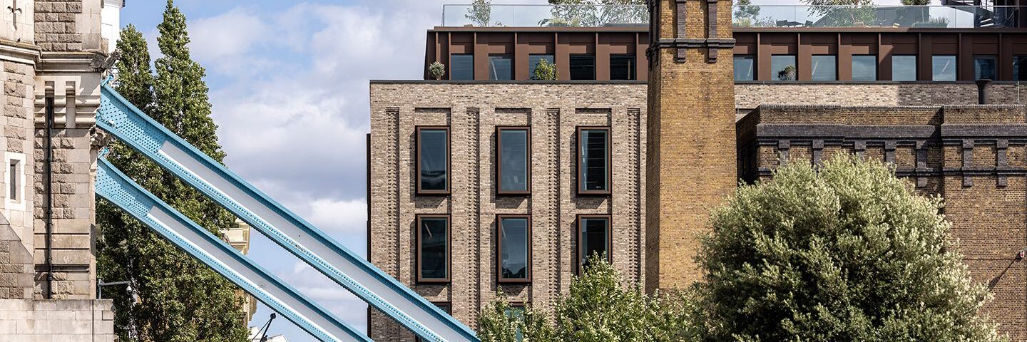Mixed-use building with brick and metal cladding details, behind a blue metal beam of the Tower Bridge.