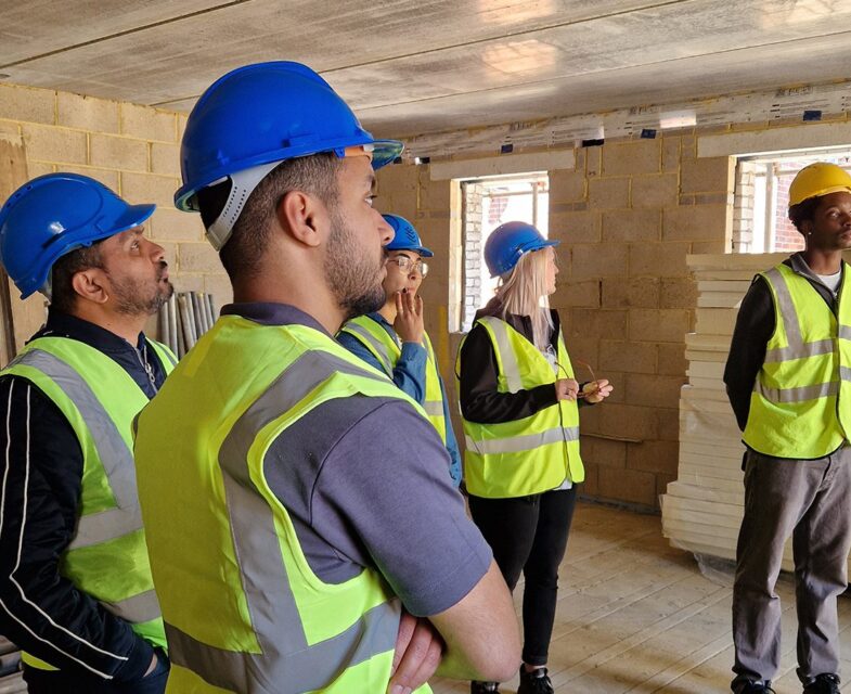 Group of five architectural staff wearing high-vis vests and hard hats looking at an unfinished building.